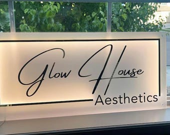 May include: A rectangular illuminated sign with the words "Glow House Aesthetics" in a stylish script font. The sign has a white background with a black border and is likely used for a business.