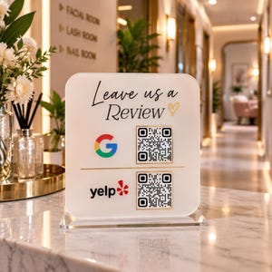 May include: A square, frosted acrylic sign with the text "Leave us a Review" and a gold heart icon. It features a Google logo and a Yelp logo, each paired with a unique QR code for scanning, mounted on a clear acrylic base.
