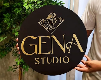 Puede incluir: Un letrero circular negro con letras doradas que dice "GENA STUDIO". Una imagen estilizada dorada de una cabeza de mujer con cabello está por encima del texto.
