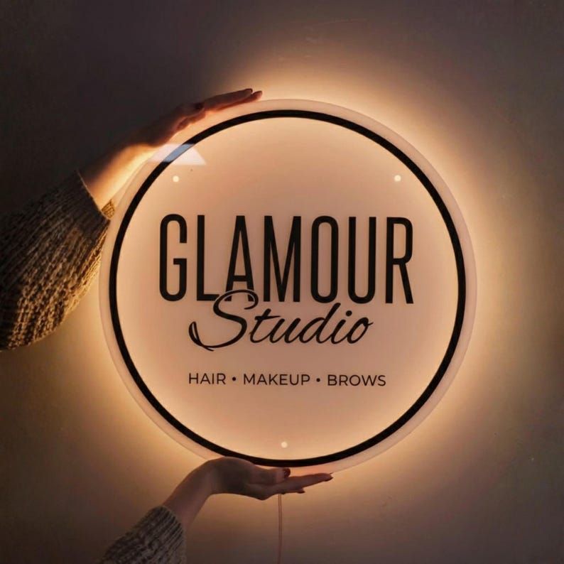 May include: A round, illuminated sign with the words "GLAMOUR Studio" in black script and block letters. The sign also includes the text "HAIR &bull; MAKEUP &bull; BROWS" in smaller font. The sign is held by two hands.