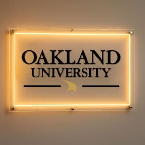 Puede incluir: Un letrero rectangular con las palabras "OAKLAND UNIVERSITY" en negro, con un logotipo amarillo y negro. El letrero tiene un borde amarillo brillante y está montado con pequeños sujetadores dorados.