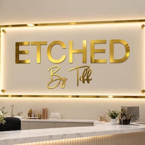 May include: A rectangular wall sign featuring gold-colored lettering that reads ETCHED By Tiff. The sign is framed in gold, mounted with four corner standoffs, and illuminated by a warm white LED backlight against a light-colored wall.