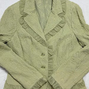 May include: A light green blazer with a textured fabric and ruffled trim. The blazer has a notched collar, three buttons, and long sleeves. The label reads "TERI JON".