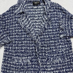 May include: A navy blue and white tweed jacket with fringe trim. The jacket has a collar and two front pockets. The fabric is woven with a textured pattern. The jacket is a stylish outerwear piece.