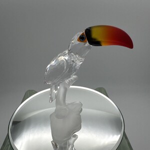 May include: A clear crystal toucan figurine perched on a frosted glass branch. The toucan has a large beak with red, yellow, and black coloring. The figurine sits on a round, mirrored surface.