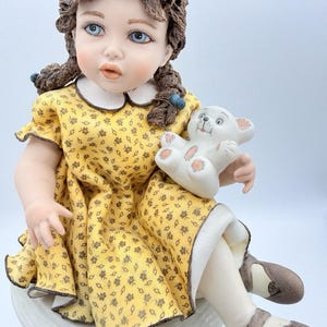 May include: A porcelain figurine of a young girl with blue eyes and brown hair in pigtails, wearing a yellow floral dress and brown shoes. She holds a white teddy bear. The figurine is seated on a white base.
