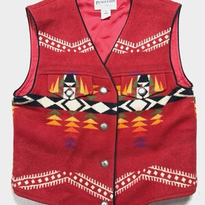 May include: A red wool vest featuring a geometric pattern in white, black, yellow, orange, and purple. The vest has a V-neckline, a front button closure with silver-toned buttons, two chest pockets with flaps, and a red lining. A Pendleton label is visible.