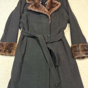 May include: A black coat with a faux fur collar and cuffs. The coat has a belted waist and two buttons down the front. The faux fur trim is brown. The coat is a classic style.