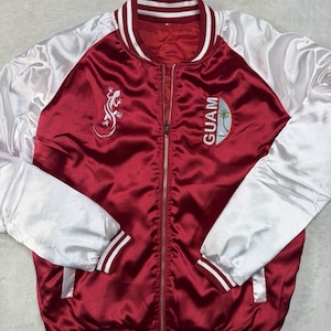 May include: A maroon and white satin bomber jacket featuring a front zipper closure, striped ribbed collar, cuffs, and hem. The chest displays an embroidered white gecko on the right and a circular patch with the text GUAM and a palm tree on the left.
