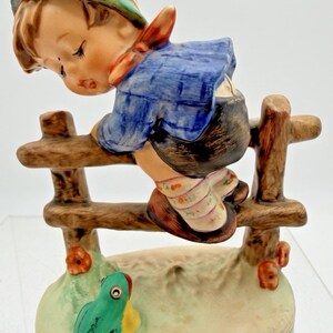 May include: A ceramic figurine of a child in a blue shirt and green hat, perched on a wooden fence. The figurine has a detailed design, with a frog at the base. The colors include brown, blue, green, and red.