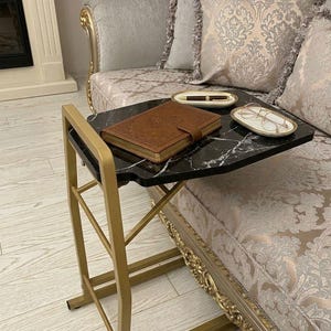 Puede incluir: Una mesa auxiliar con tapa de mármol negro y estructura metálica dorada. La mesa tiene un libro encuadernado en cuero marrón, un bolígrafo y gafas en una bandeja decorativa. La mesa está junto a un sofá con cojines estampados.