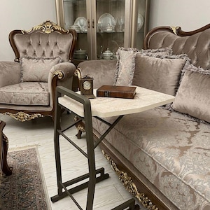 Puede incluir: Una escena de salón con muebles ornamentados. Una pequeña mesa auxiliar con tapa de mármol y estructura de metal oscuro sostiene un libro y un reloj. El sofá y el sillón están tapizados en una tela estampada con detalles dorados, creando un ambiente lujoso.