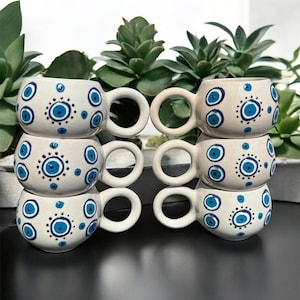 May include: A set of six ceramic mugs with a cream-colored base and white handles. Each mug is decorated with blue eye-shaped designs. The mugs are stacked in pairs, with green plants in the background.