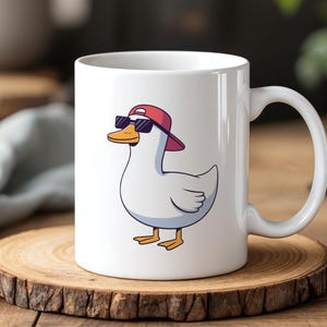May include: White ceramic mug featuring a cartoon duck wearing a red cap and sunglasses. The duck has a yellow beak and feet, with a blue outline. The mug is on a wooden coaster.