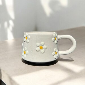 May include: A white ceramic mug with a daisy pattern. The mug has a handle and is decorated with yellow and white daisies.