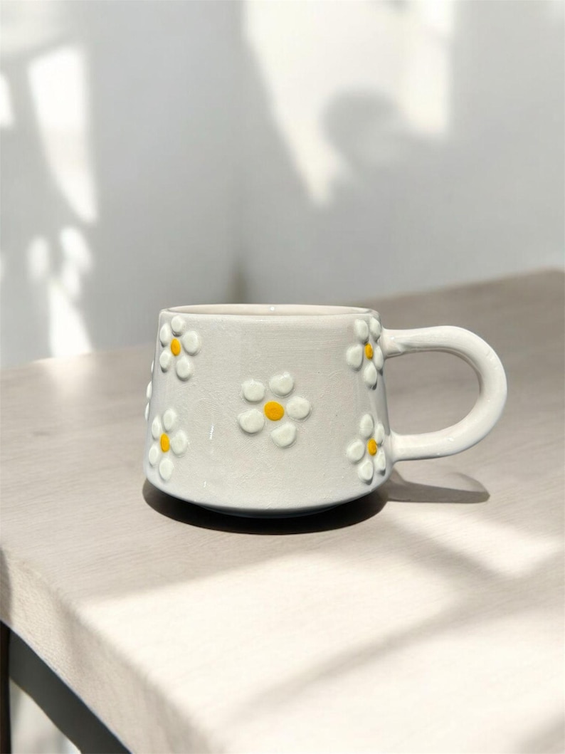 May include: A white ceramic mug with a daisy pattern. The mug has a handle and is decorated with yellow and white daisies.