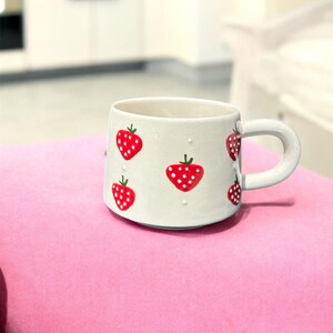 May include: A white ceramic mug with a rounded handle, adorned with red strawberries. Each strawberry has white polka dots and green stems. The mug is set on a pink surface.