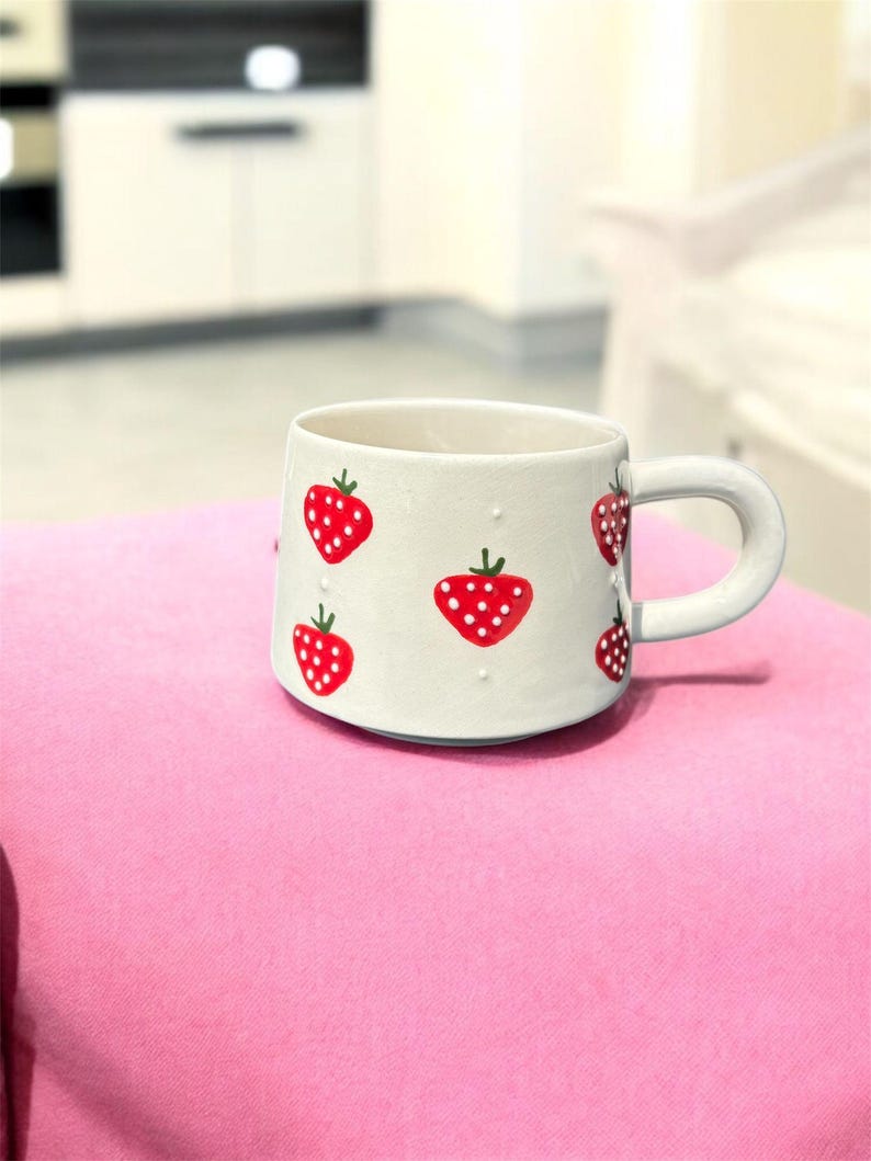 May include: A white ceramic mug with a rounded handle, adorned with red strawberries. Each strawberry has white polka dots and green stems. The mug is set on a pink surface.