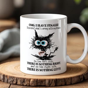 May include: White ceramic mug with a humorous black cat design and text. The cat has large blue eyes and holds a coffee cup. The mug's text reads, "OMG. I HAVE FINALLY Discovered what's wrong with my brain: On the left side, THERE IS NOTHING RIGHT..."