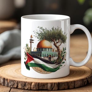 Pode incluir: Caneca de cerâmica branca com uma ilustração detalhada de um marco do Médio Oriente com uma cúpula dourada, um minarete e a bandeira palestina. O design inclui ramos de oliveira e uma árvore estilizada. A caneca está sobre um descanso de madeira.
