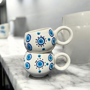 Może przedstawiać: Dwa białe ceramiczne kubki ułożone jeden na drugim, każdy ozdobiony niebiesko-czarnymi wzorami w kształcie oka. Kubki mają zaokrąglone uchwyty i stoją na marmurowej powierzchni. Biały dzbanek w tle.