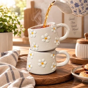May include: Two stacked ceramic mugs with a light gray finish and raised white daisy patterns featuring yellow centers. A floral patterned teapot pours steaming amber tea into the top mug. The mugs have rounded handles and a smooth, glossy texture.