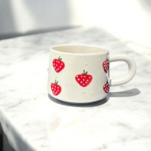 May include: A white ceramic mug decorated with red strawberries. The strawberries have white dotted seeds and green stems. The mug has a rounded handle and sits on a white marble surface.