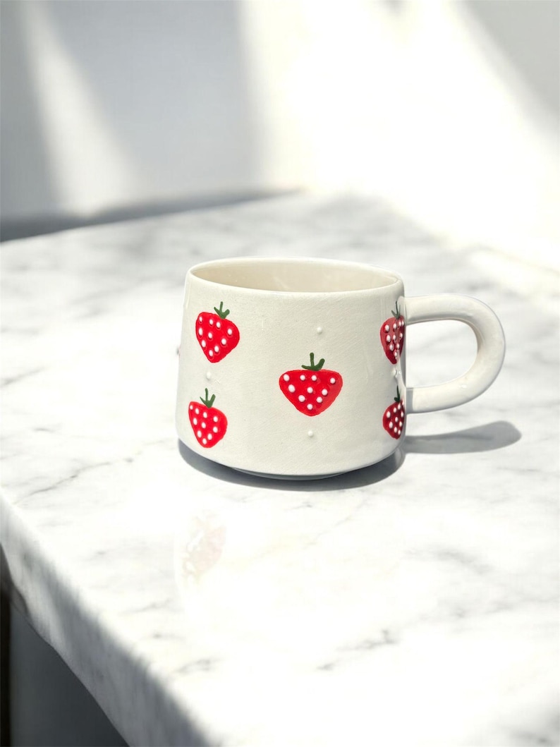 May include: A white ceramic mug decorated with red strawberries. The strawberries have white dotted seeds and green stems. The mug has a rounded handle and sits on a white marble surface.