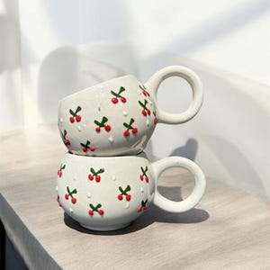 Może przedstawiać: Dwa białe ceramiczne kubki ułożone jeden na drugim. Każdy kubek ma okrągły korpus, okrągły uchwyt i jest ozdobiony czerwonymi i zielonymi wzorami wiśni. Kubki stoją na jasnej drewnianej powierzchni. Wysokość ok. 10 cm.