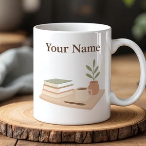 Könnte beinhalten: Weiße Keramiktasse mit dem Text "Your Name" in Braun. Die Tasse zeigt Illustrationen von Büchern, einer Kaffeetasse, einer Pflanze und einer kleinen Vase. Die Tasse steht auf einem Holzuntersetzer.