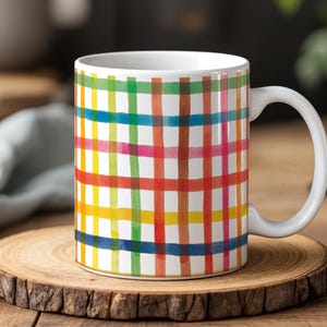 May include: White ceramic mug with a colorful plaid pattern in shades of red, orange, yellow, green, blue, and pink. The mug has a white handle and is sitting on a wooden coaster.