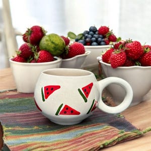 Pode incluir: Caneca de cerâmica branca com um padrão de melancia. A caneca tem uma alça e está sentada em um descanso de mesa listrado.
