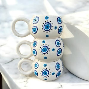 May include: Three stacked white ceramic mugs with large handles. Each mug is decorated with a blue eye pattern, featuring a central blue circle surrounded by smaller blue circles and black dots. The mugs are stacked on a white marble surface.