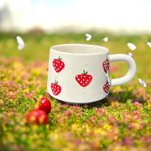 May include: A white ceramic mug with a curved handle, decorated with red strawberries. The strawberries have white polka dots and green stems. The mug is set on a bed of green and pink flowers.