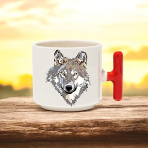 May include: White ceramic mug featuring a detailed illustration of a wolf's face in shades of gray, brown, and tan. The mug has a bright red handle. The mug is sitting on a wooden surface.