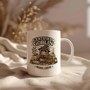 Könnte beinhalten: Ein weißer Keramikbecher mit einer Grafik eines Waschbären, der einen Zaubererhut trägt und eine kleine Tasse hält. Der Text lautet CERTIFIED GOBLIN und BOOKS, COFFEE. Die Illustration zeigt eine Topfpflanze, ein offenes Buch und Fläschchen.