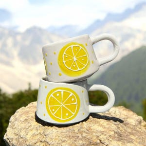 Könnte beinhalten: Zwei weiße Keramikbecher, die auf einem Felsen gestapelt sind. Jeder Becher hat ein gelbes Zitronenscheibendesign mit weißem Umriss und gelben Punkten. Die Becher haben weiße Griffe.