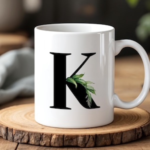 Può includere: Tazza in ceramica bianca con una grande lettera "K" nera decorata con dettagli botanici verdi. La tazza è appoggiata su un sottobicchiere di legno, un design semplice ed elegante.