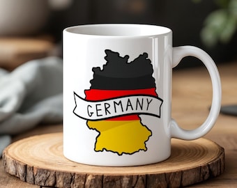 May include: White ceramic mug featuring a stylized map of Germany in black, red, and yellow, with the word "GERMANY" on a banner. The mug is on a wooden coaster.