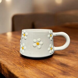 May include: A white ceramic mug with a daisy pattern. The mug has a handle and is decorated with yellow and white daisies.