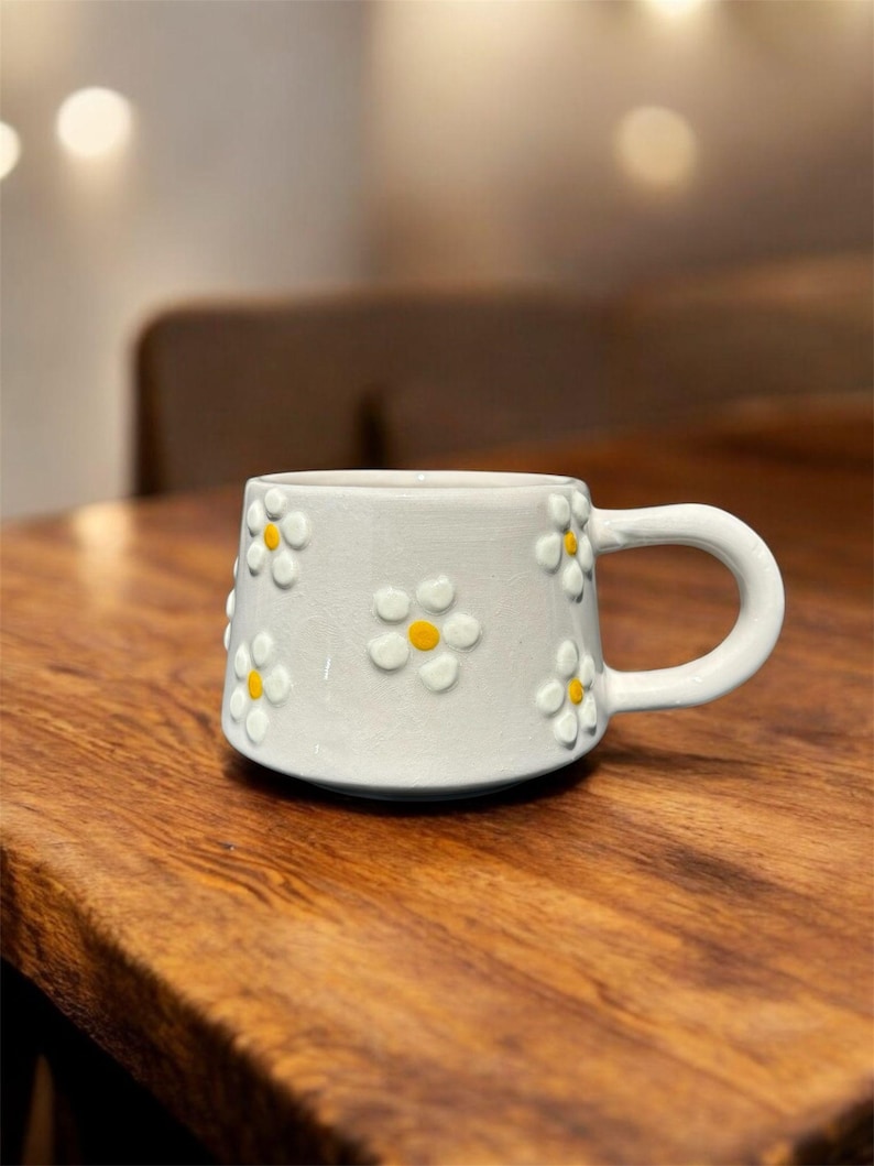May include: A white ceramic mug with a daisy pattern. The mug has a handle and is decorated with yellow and white daisies.