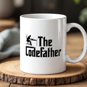 Può includere: Tazza da caffè in ceramica bianca con una grafica nera di una persona che lavora su un laptop e le parole "The Codefather". La tazza è su un sottobicchiere di legno.