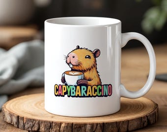 Op de afbeelding: Witte keramische mok met een cartoon capibara die een koffiekopje vasthoudt. De mok heeft het woord "CAPYBARACCINO" in kleurrijke letters. De mok staat op een houten onderzetter.