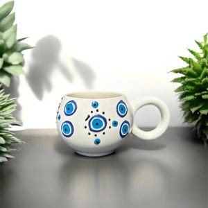 May include: White ceramic mug with a blue and black evil eye design. The mug has a rounded body and a handle.