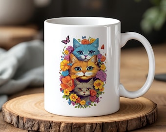 May include: White ceramic mug featuring a colorful illustration of three cats surrounded by flowers and butterflies. The cats are blue, orange, and gray, with vibrant floral accents in orange, yellow, and purple. The mug has a curved handle.
