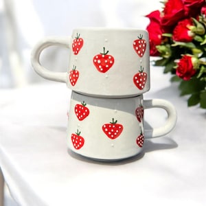 Può includere: Due tazze in ceramica bianca impilate, decorate con un motivo ripetuto di fragole rosse con pois bianchi. Le tazze hanno manici curvi e una finitura liscia. Perfette per caffè o tè, queste tazze aggiungono fascino.