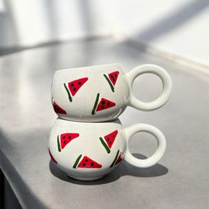 Könnte beinhalten: Zwei weiße Keramikbecher, die übereinander gestapelt sind. Jeder Becher ist mit einem sich wiederholenden Muster aus roten Wassermelonenscheiben mit grünen Schalen verziert. Die Becher haben große, runde Griffe und eine glänzende Oberfläche. Die Becher stehen auf einer hellgrauen Oberfläche.