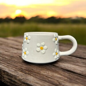 May include: White ceramic mug with a daisy pattern. The mug has a handle and is sitting on a wooden surface.