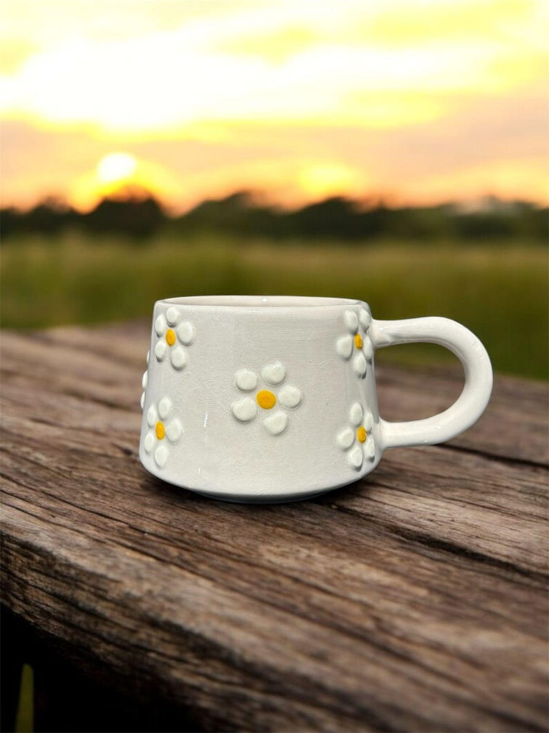 May include: White ceramic mug with a daisy pattern. The mug has a handle and is sitting on a wooden surface.