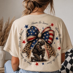 Könnte beinhalten: Beige T-Shirt mit patriotischem Design. Auf der Rückseite des Shirts befindet sich eine Schleife im Stil der amerikanischen Flagge, ein kniender Soldat und der Text "Honor the brave" in kreisförmiger Anordnung. Rote und blaue Sterne ergänzen das Design.